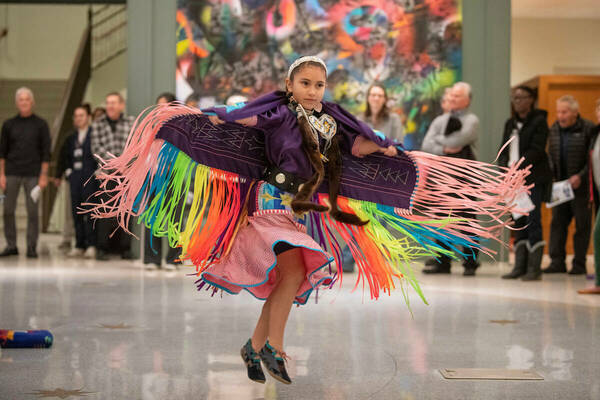 People look on as an Indigenous youngster dances and her colorful regalia is suspended in air.