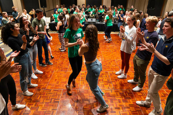 Two women dance on a parquet floor while participants clap.