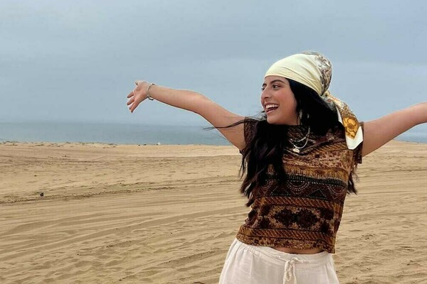 A woman wearing a bandana on her head and standing on sand smiles and extends her arms out from her sides.