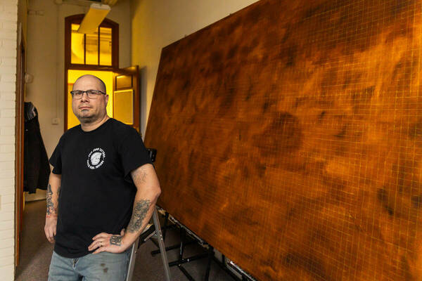 David Martin, wearing glasses and a black T-shirt, stands with his hands on his hips looking at the camera. His large brown painting-in -progress is behind him.
