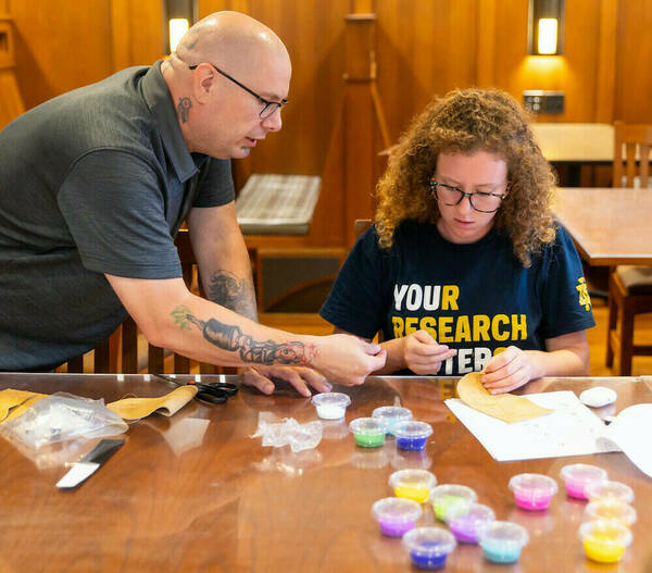 A man with glasses and wearing a dark gray T-shirt leans over to give instruction to a student working on a beading project. Small plastic cups with a variety of colored beads are on the table.