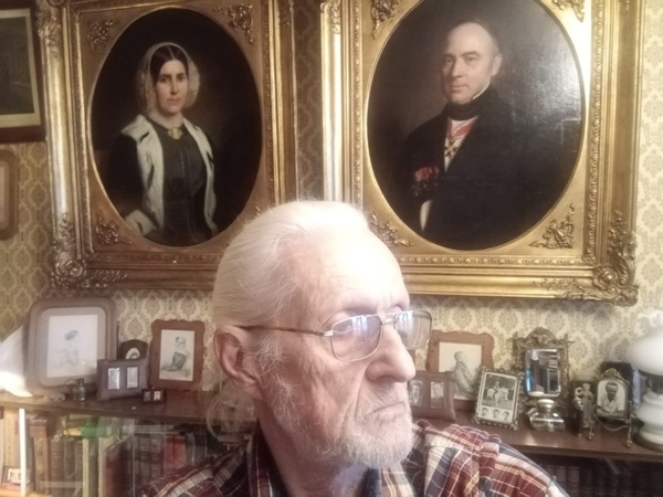 A man with white hair and glasses is in profile. Portraits of his ancestors are on the wall behind him in large golden frames.