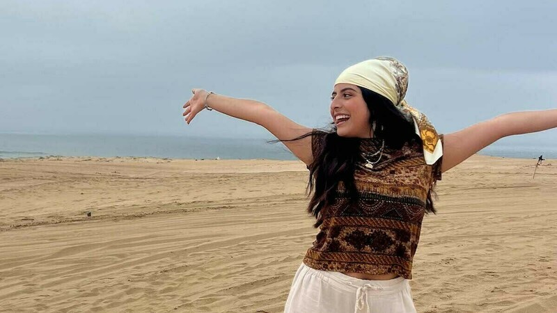 A woman wearing a bandana on her head and standing on sand smiles and extends her arms out from her sides.