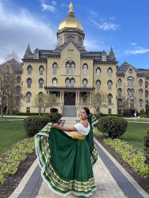A woman wearing a traditional dance dress poses in front of the Main Building at Notre Dame.