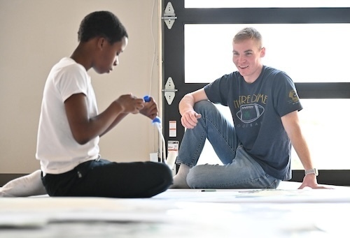 A youth in a shite T-shirt and a Notre Dame student in a blue T-shirt sit on the floor.