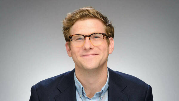 A man with brown hair and glasses, wearing a dark blue blazer.