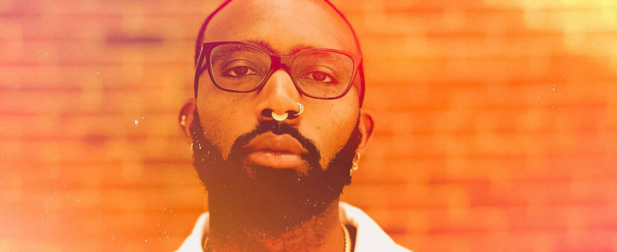 Joseph Earl Thomas, presenting as a black male with a beard, glasses, and nose piercings. He is standing in front of a brick wall and the photo has a orange-sepia filter added.