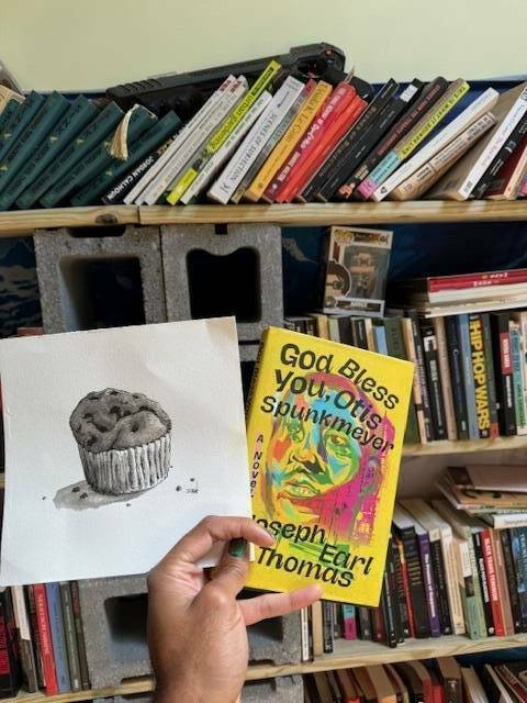 A hand holding a sketch drawing of a muffin, and the colorful cover of the book God Bless You, Otis Spunkmeyer by Joseph Earl Thomas.