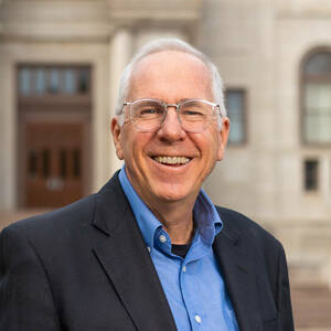 A smiling man with short white hair and glasses, wearing a blue shirt and dark blazer, stands before the stone entrance, columns, and steps of the Notre Dame Main Building.