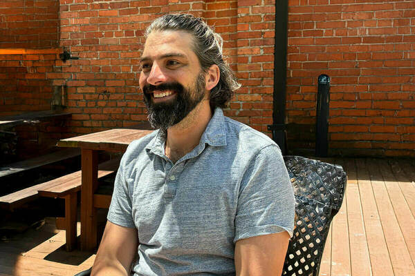 A man with a beard and graying hair pulled back smiles as he sits on a black metal chair on a wooden deck. He wears a gray polo shirt. Behind him is a brick wall and wooden picnic tables.