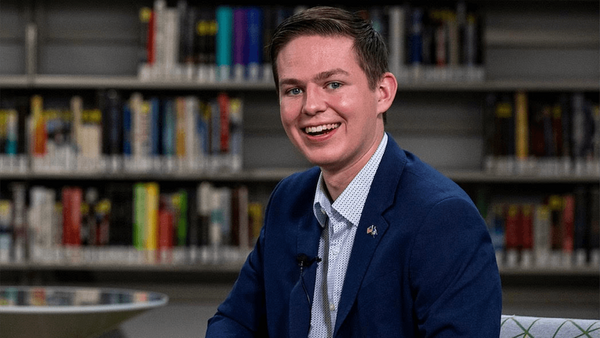 A smiling young man in a navy blue blazer and patterned white shirt sits in front of library bookshelves.  A small lavalier microphone is clipped to his lapel.