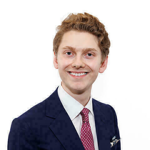 Headshot of a light-skinned young man with wavy red hair, smiling, wearing a navy suit, white shirt, and maroon patterned tie against a white background.
