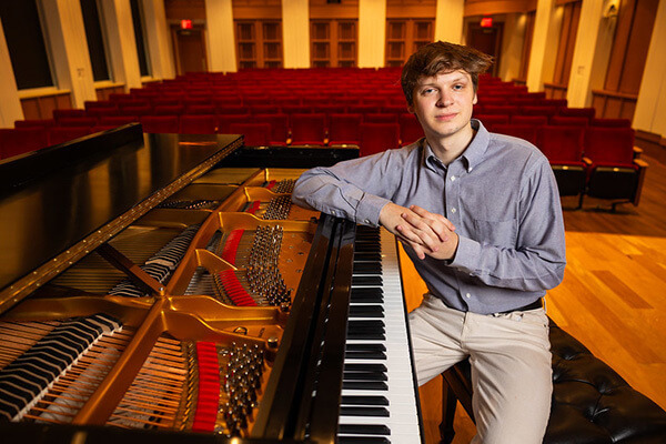 A young man in a blue shirt and khaki pants sits with his hands clasped, leaning against a grand piano on a stage with red seats in the audience.