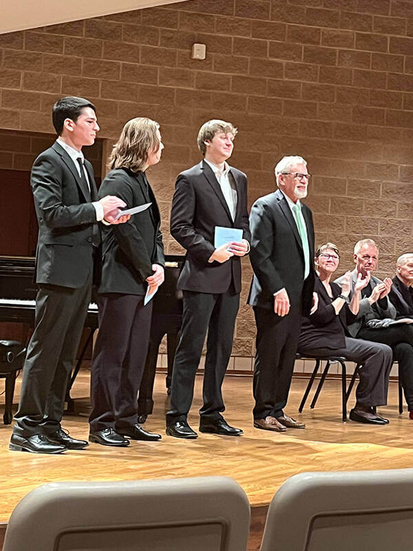 Three young men in suits stand on a stage with a man in a suit and green tie, as audience members applaud.