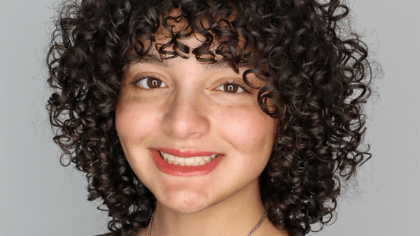 Headshot of a young person with tight, dark curly hair, brown eyes, and a broad smile against a plain gray background.