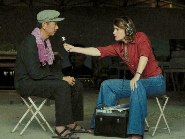 Woman in red shirt interviewing man in a green hat, black outfit, and black sandals.