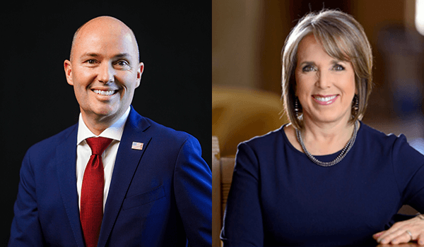 On the left is Spencer Cox, a smiling bald man in a navy blue suit, white shirt, and red tie. On the right is Michele Lujan Grisham, a smiling woman with short light brown hair wearing a navy blue top and a necklace.