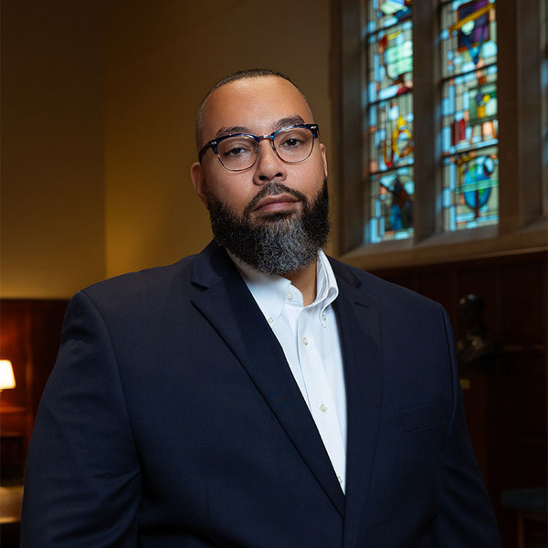 Headshot of a person with glasses and a dark beard in front of a brick wall.  They are wearing a maroon turtleneck and a light brown jacket.