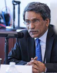 A man with graying hair and glasses is speaking into a microphone at a conference or meeting. He is wearing a dark suit, light blue shirt, and a patterned blue tie. His hands are clasped in front of him on the table. There are blurred office items and people in the background.