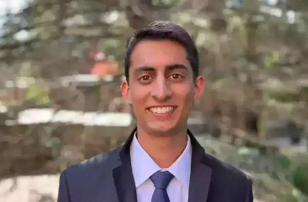 Smiling young man with dark hair and brown eyes in a dark suit, white shirt, and blue patterned tie. Blurred green and brown foliage in the background.
