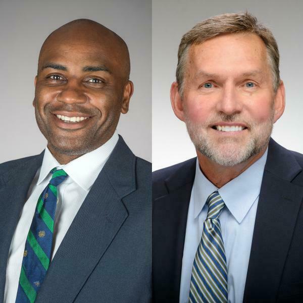 Two smiling men in suits against a gray background. The man on the left is bald with dark skin, wearing a blue suit and a green and blue striped tie with gold shamrocks. The man on the right has a gray beard and short brown hair, wearing a blue suit and a striped blue and green tie.