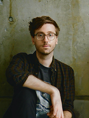 A person wearing dark clothing and glasses plays a black keyboard. The scene is bathed in red light, with a bright white light tube angled in the upper left.