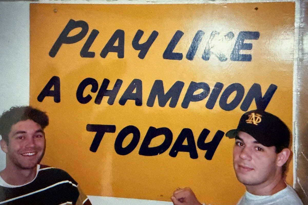 Two smiling young men pose in front of the yellow "Play Like a Champion Today" sign. One wears a striped shirt, the other a navy baseball cap with a gold ND logo.