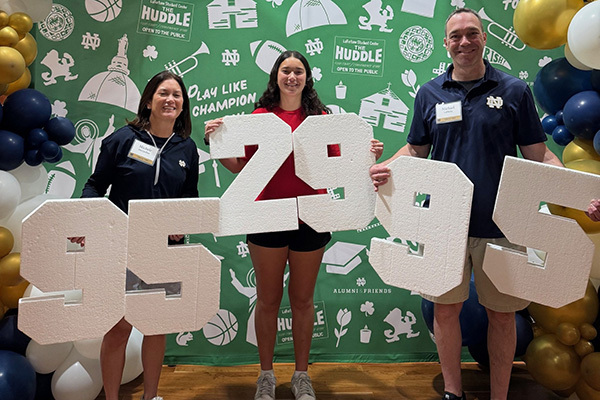 Three smiling people stand against a green Notre Dame-themed backdrop. A woman in a dark blue top and another in a red shirt hold "95" and "29", respectively. A man in a dark blue polo holds "95". Together, they display "952995". Gold, blue, and white balloons are on the sides.