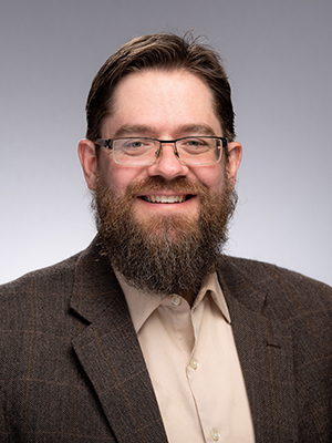 A smiling man with a full brown beard, dark-rimmed glasses, and a brown patterned jacket over a light collared shirt.