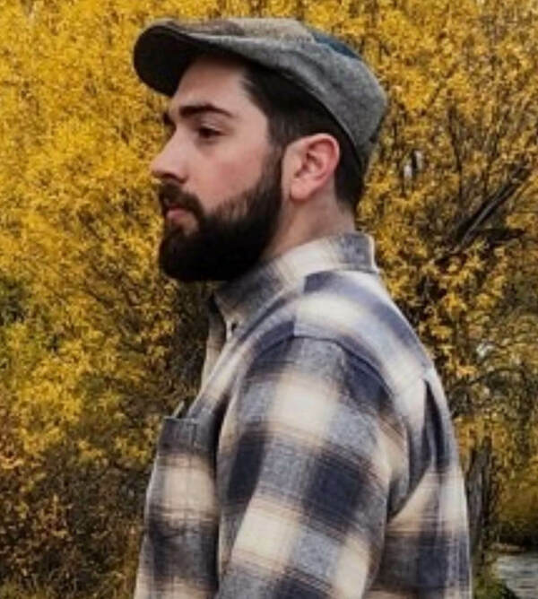Man in grey flat cap, blue-beige plaid shirt, and beard, looking left. Vibrant yellow autumn foliage in background.