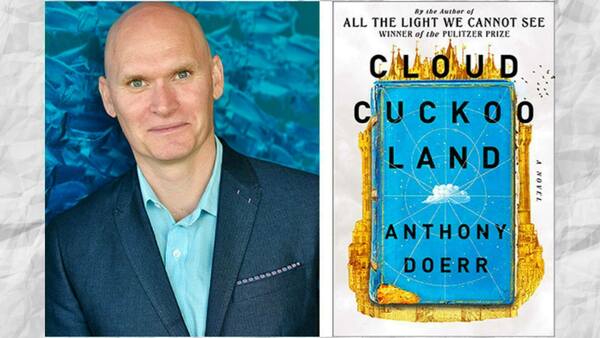 Bald Pulitzer Prize winner Anthony Doerr smiles in a blue suit, beside his novel Cloud Cuckoo Land. Blue and gold cover.