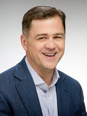 A headshot of a man with short brown hair, wearing a textured navy blazer and a light purple and white checked collared shirt. He is smiling broadly against a plain gray background.