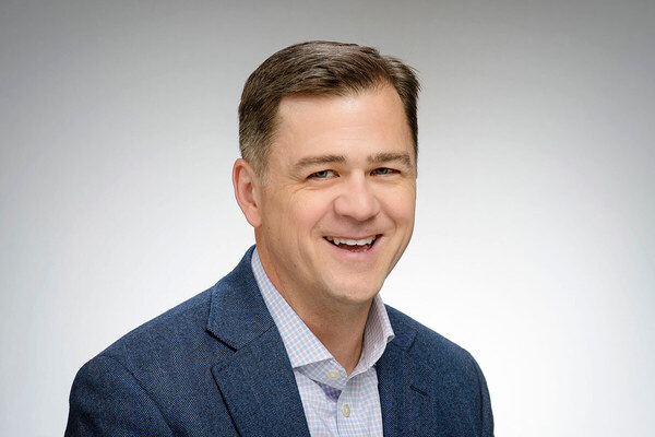 A headshot of a man with short brown hair, wearing a textured navy blazer and a light purple and white checked collared shirt. He is smiling broadly against a plain gray background.