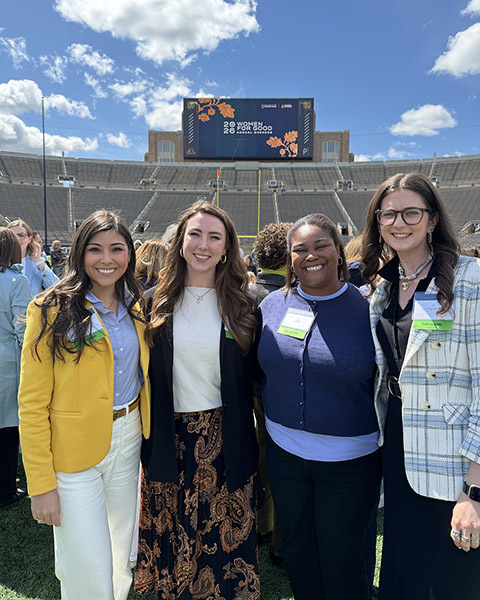Four smiling women on Notre Dame Stadium field. Scoreboard shows '20 WOMEN 26 FOR GOOD' beneath a blue sky.