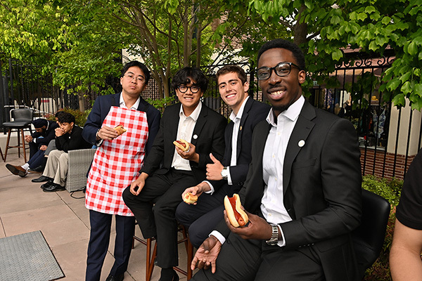 Four young men in suits, one with a red and white checkered apron, smile at the camera while holding hot dogs.
