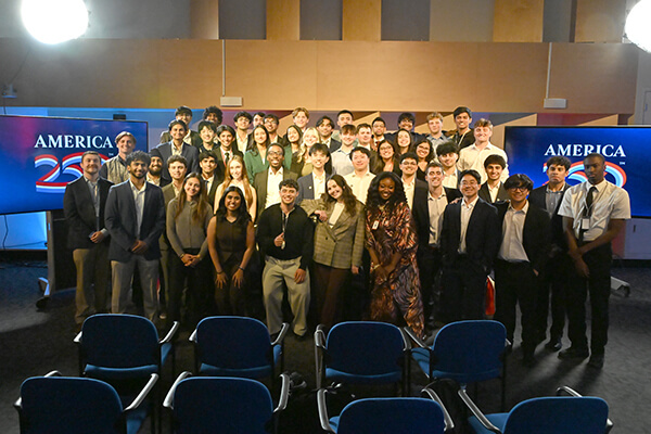 A group photo of four rows of people in formal wear in a meeting room with chairs in the foreground and screens reading America 250 to the sides in the background.