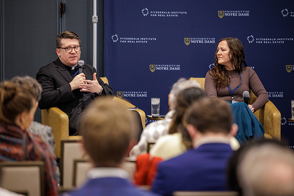 A man wearing glasses and a priest's black outfit with a white clerical collar speaks into a microphone, sitting on a small stage in front of a seated audience. To his right is a woman with long brown hair also sitting on the stage, listening to him talk.