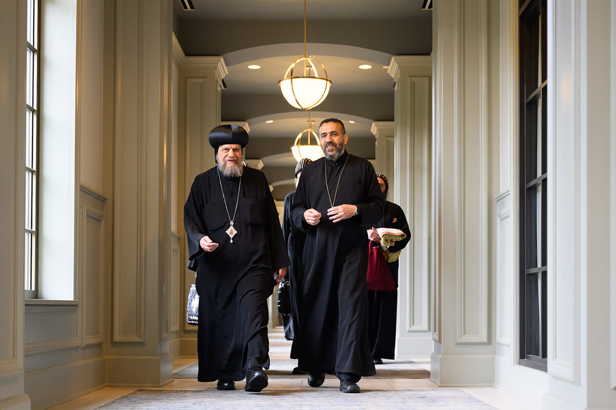 Two bearded clergy in black robes, the one on the left wearing a round black hat called an emma, walk smiling down a bright hallway with white fluted columns.