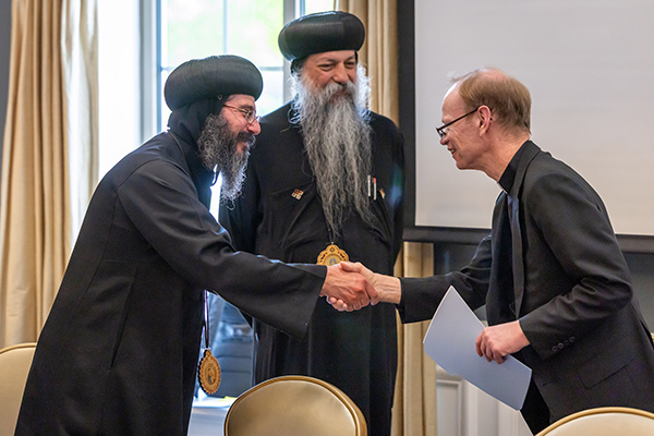 Bearded man in black robes and head covering shakes hands with a man in a suit as another bearded man smiles.