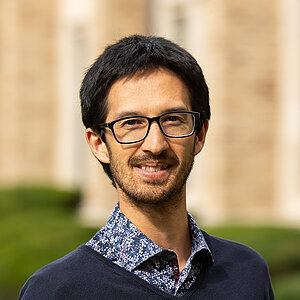 Man wearing glasses and a dark sweater over a patterned shirt smiles in front of a tan building.