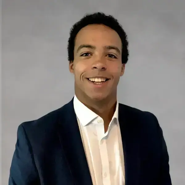 Man with dark curly hair, wide smile, wearing navy suit jacket and white collared shirt on gray background.