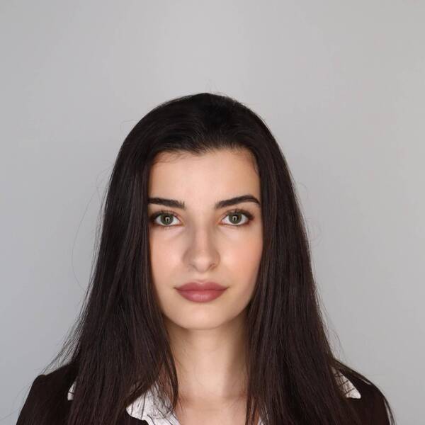 Woman with long, straight, dark hair wearing a black suit jacket and a striped collared shirt with a gray background