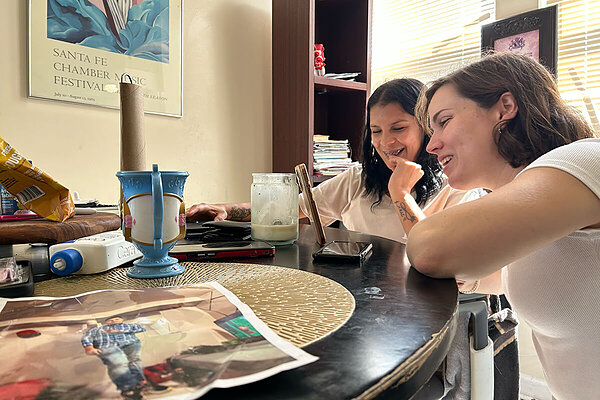 Two smiling women, one with dark hair, the other with brown hair, look right and smile at a phone propped up on a table.