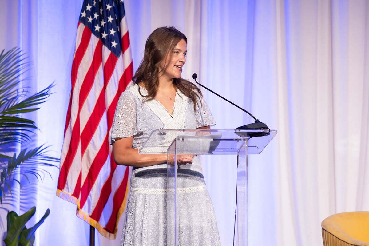 Woman in a light blue dress speaks at a podium next to an American flag.