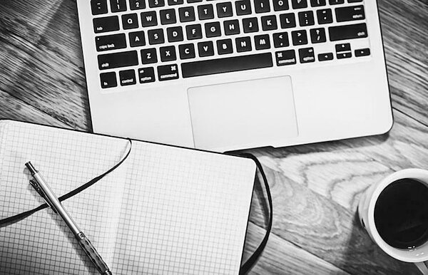 Black and white photo of a laptop keyboard, a gridded notebook with a pen, and a cup of coffee on a wooden desk.