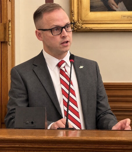 Man in a grey suit and red and white striped tie speaks at a podium.
