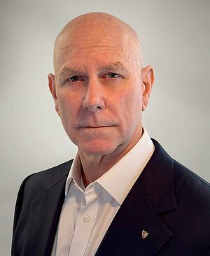 Bald man in a suit and white shirt with a crest pin.